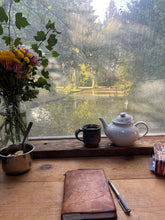 Tea set with a teapot, cup, and notebook on a wooden surface with a window view of trees.